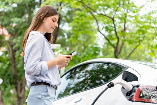 Young woman using smartphone online banking application to pay for electric car battery charging from EV charging station during vacation holiday road trip at national park or summer forest. Exalt