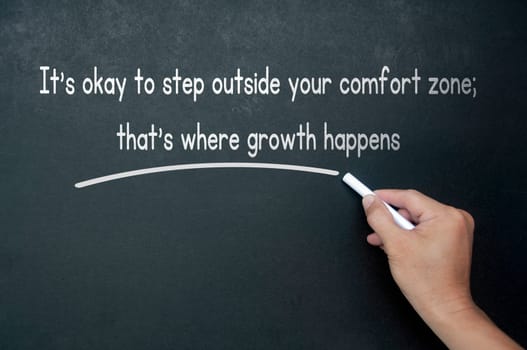 Hand writing It is okay to step outside your comfort zone affirmation on black board.