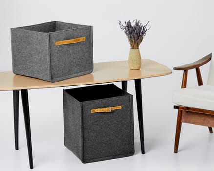 Modern minimalist interior with two grey felt storage boxes on sleek desk, complemented by wooden armchair and vase with lavender