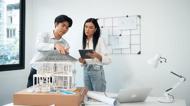 Beautiful young caucasian project manager tells asian engineer about building detail by using tablet while engineer measuring house model on meeting table with blueprint scatter around. Immaculate