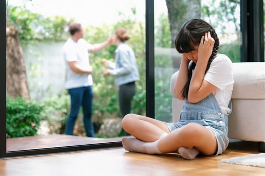 Stressed and unhappy young girl huddle in corner, cover her ears blocking sound of her parent arguing in background. Domestic violence at home and traumatic childhood develop to depression. Synchronos