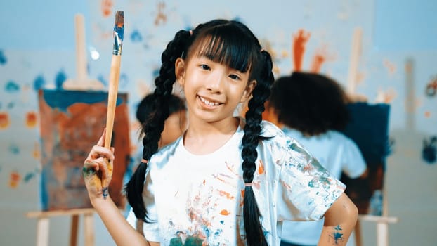 Creative cute girl pose at camera while diverse children painting behind. Young beautiful child looking at camera while standing at stained room and holding paintbrush. Creative activity. Erudition.