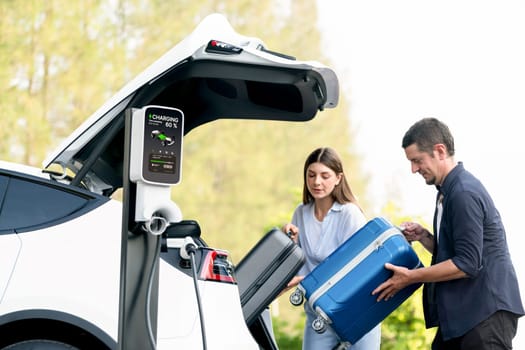 Lovely young couple recharging battery for electric car while relaxing and sitting on the trunk during road trip travel EV car in autumnal forest. Eco friendly travel on vacation during autumn. Exalt