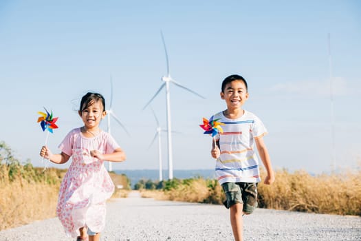 Brother and sister run by windmills holding pinwheels. Kids' joy near turbines represents a future of clean energy. Smiling children carefree and happy engage with sustainable technology.
