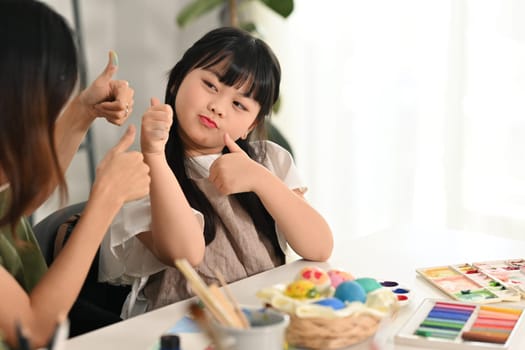 Playful little child girl painting Easter eggs with mother in living room. Preparing for Easter concept.