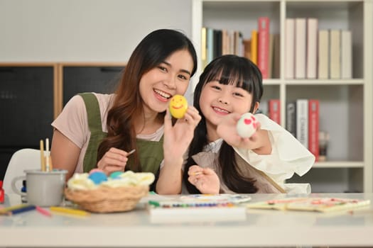 Happy mother and her daughter preparing for Easter painting eggs in living room.