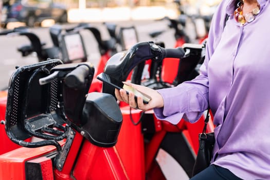 detail of the hand of a senior woman unlocking rental bike with the app on her mobile phone, concept of sustainable mobility and active lifestyle in elderly people