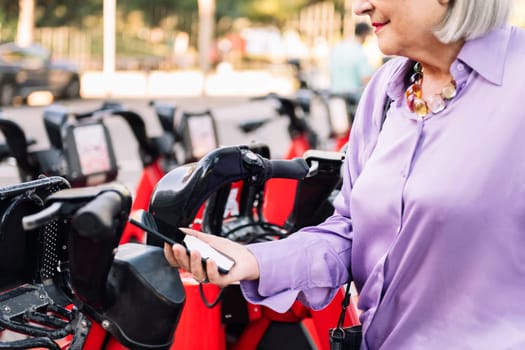 unrecognizable senior woman unlocking rental bike with the app on her mobile phone, concept of sustainable mobility and active lifestyle in elderly people