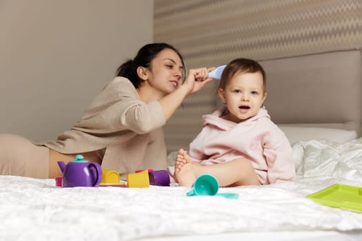 Happy mother is brushing her cute little daughter hair on bed