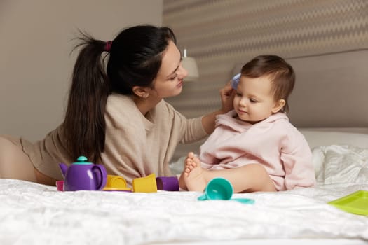 Happy brunette mother is combing her cute little daughter hair on bed