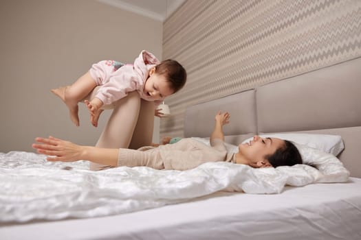 Happy Caucasian mother raising laughing cute baby daughter and playing together in bedroom