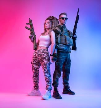 A daring stylish girl with an automatic rifle and a guy in military clothes with an airsoft gun in neon light on a bright neon background pose fashionably