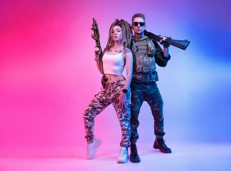 A daring stylish girl with an automatic rifle and a guy in military clothes with an airsoft gun in neon light on a bright neon background pose fashionably