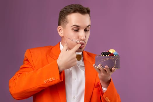 handsome birthday man in orange jacket eating cake on purple background