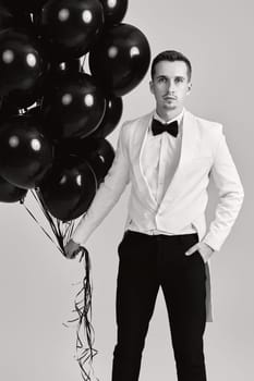 elegant caucasian man in white suit tuxedo with bunch of black air balloons on white background.