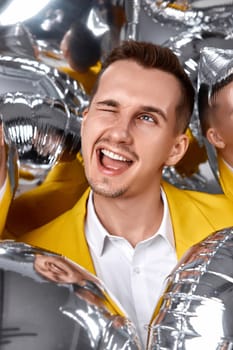 happy winking man in yellow jacket with a lot of silver air balloons. birthday party