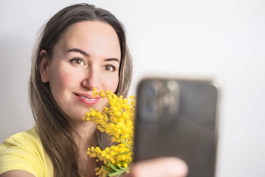 Beautiful young woman with mimosa flowers taking a selfie on white background.