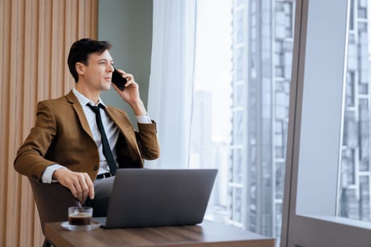 Closeup of handsome businessman making phone call with manager while sitting near window with skyscraper view. Executive manager talking working by using phone and laptop. Look aside. Ornamented.