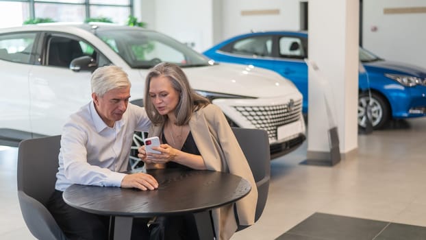 A mature couple is sitting in a car dealership and looking at a smartphone