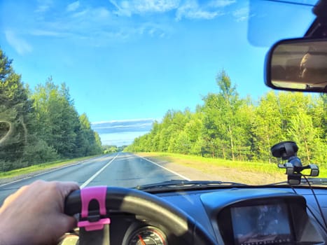 Car salon, windshield, hand of woman on steering wheel and landscape. View from seat of driver on nature with Road, trees, blue sky at a sunny day. Single trip of female traveler. Partial focus