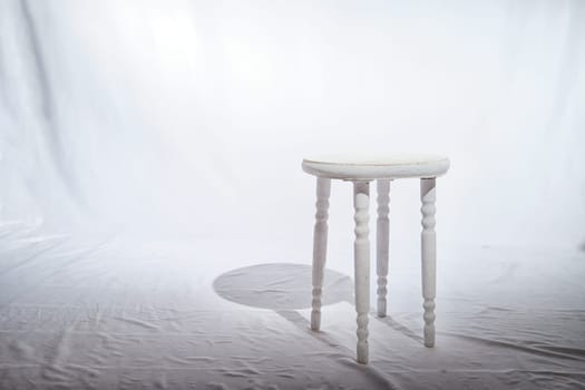 Solitary Wooden Stool in a White Minimalist Setting. Lone, white-painted wooden stool stands centered on textured surface, surrounded by soft white backdrop