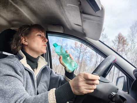 Portrait of female driver in solo journey. Adult mature woman holding steering wheel and bottle of water. Drinking while driving for to stay awake. Lady girl drinks alcohol vodka or gin while driving