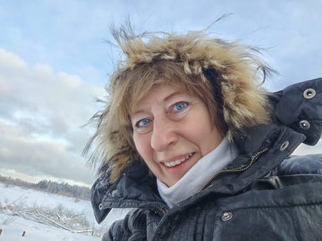 Happy, cheerful middle-aged woman in a warm jacket and a hood with fur taking selfie in winter outdoors. Female Traveler in Alaska, Lapland, Arctic, Siberia