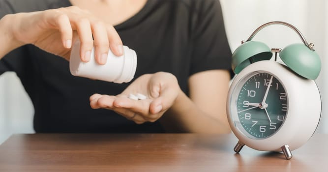 Person in a black t-shirt pouring pills into the hand with an alarm clock on the table for time management, deadlines, and the concept of medical and health care