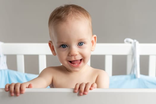 Baby girl with blue eyes standing in crib holding rail and smiling
