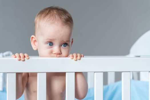 Sad and lonely baby girl standing in a white wooden crib, holding and biting the rail with a sad