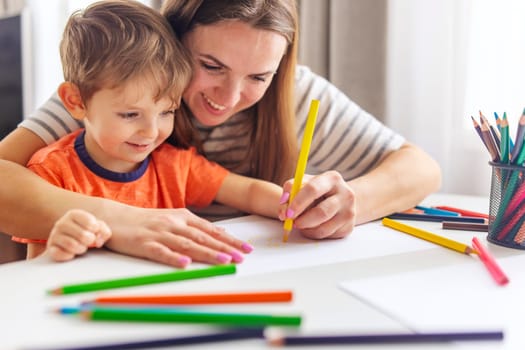 A young boy grins broadly as his mother assists him in drawing with pencils, enjoying a creative afternoon together.