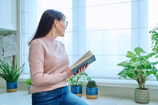 Middle-aged woman reading a fiction book at home, at home near the window, looking out the window thoughtfully, winter season