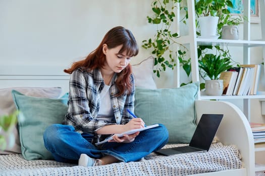 College student girl studying at home, sitting on the couch with laptop, making notes in study book. Education, training, technology, youth concept