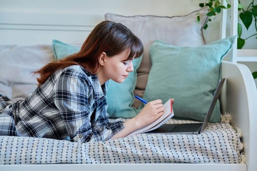 College student girl studying at home, lying on the couch with laptop, making notes in study book. Education, training, technology, youth concept