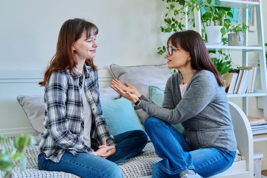 Talking happy smiling mother and teenage daughter sitting together on couch at home. Family, communication, motherhood, friendship, relationship between parent and daughter 18-20 years old