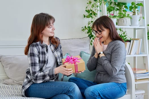 Cheerful teenage daughter congratulating middle aged mother with gift, sitting together on couch at home. Holiday birthday, congratulations, love, mother's day, happiness joy concept