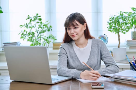 College student girl sitting at desk using laptop computer, making notes in study notebook, at home. Teenager female watching webinar, preparing for exam tests, studying remotely.