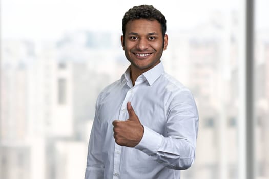 Smiling indian businessman looking at camera and giving thumb up gesture. Young smart man gesturing thumb up on blurred background.