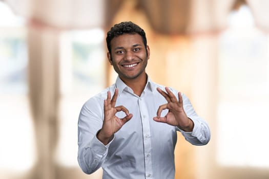 Smiling businessman wearing white shirt showing okey symbol with both hands. Blur interior background.