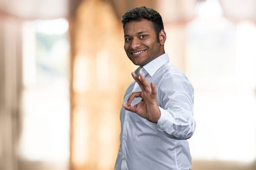 Attractive young businessman looking at camera and showing okey symbol. Blur interior background.