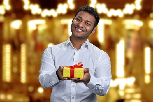 Successful Indian businessman offering gift box on golden bokeh background. Present for Christmas holiday.