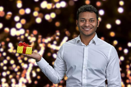 Smiling handsome man in white shirt holding gift box. Christmas bokeh lights in the background.