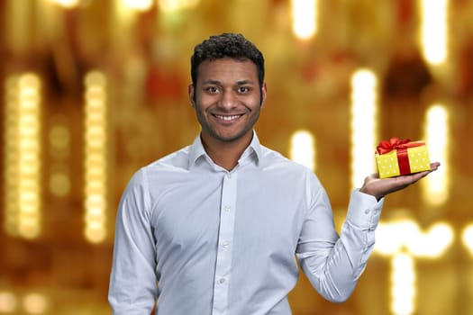 Young smiling man holding gift box on his palm and looking at camera. Time for Christmas gifts.