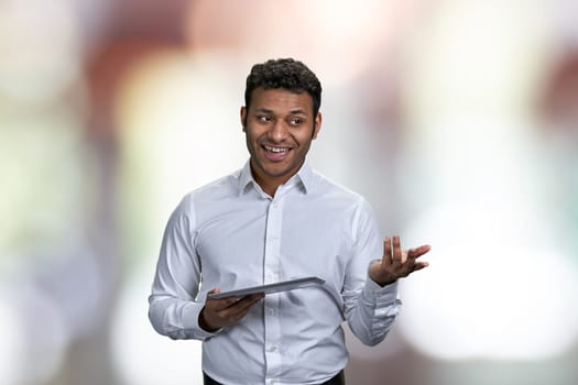 Cheerful young business coach talking against abstract bokeh background. Businessman speaker using digital tablet.