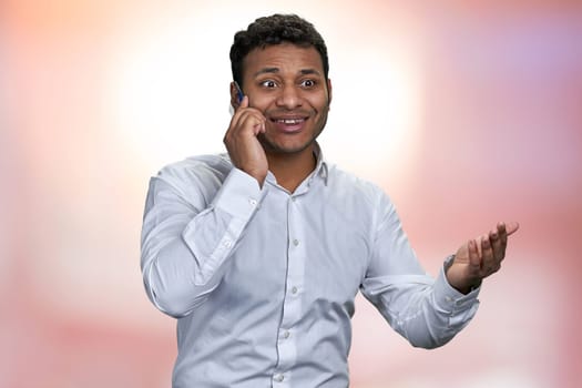 Delighted businessman talking on mobile phone on pink abstract bokeh background. Excited indian man talks on cell phone.