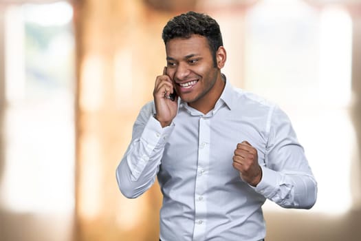 Excited young indian businessman talking on cell phone. Happy businessman talks on mobile phone on blurred background.
