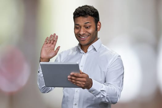 Happy young businessman waving while having video call on digital tablet. Abstract bokeh background.