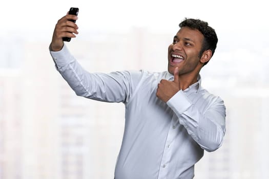 Cheerful asian businessman giving thumb up while taking selfie with smartphone. Happy indian man in white shirt using cell phone on blurred background.