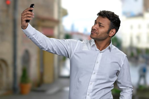 Young handsome man taking selfie with smartphone on blur city street background. People, technology and lifestyle concept.