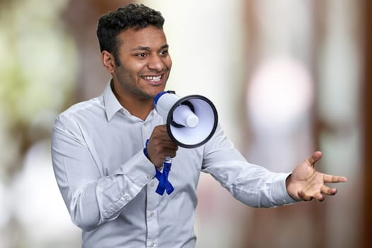 Cheerful young man in white shirt talking into megaphone. Abstract bokeh background.